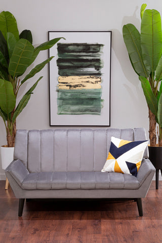 A grey two-seat sofa with a tufted design, positioned in a modern living space. The sofa features black legs and is adorned with a colorful geometric throw pillow. Behind the sofa, there is a large framed abstract artwork and two tall green plants, creating a stylish and inviting atmosphere. The floor is made of warm wood, adding to the cozy ambiance