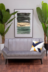 A grey two-seat sofa with a tufted design, positioned in a modern living space. The sofa features black legs and is adorned with a colorful geometric throw pillow. Behind the sofa, there is a large framed abstract artwork and two tall green plants, creating a stylish and inviting atmosphere. The floor is made of warm wood, adding to the cozy ambiance