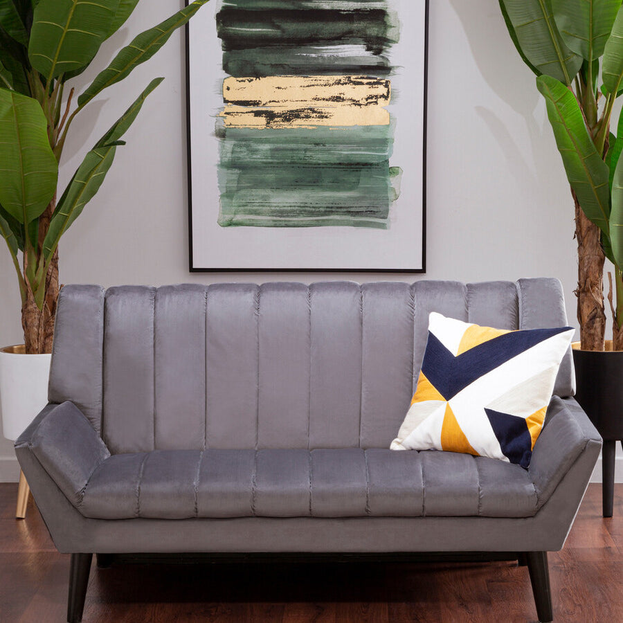 Two-seat grey sofa with a modern design, featuring vertical stitching on the backrest and tapered wooden legs. A decorative pillow with a geometric pattern in yellow, navy, and white is placed on one side. In the background, there is a large abstract painting and two tall green plants, set against a light-colored wall and wooden floor
