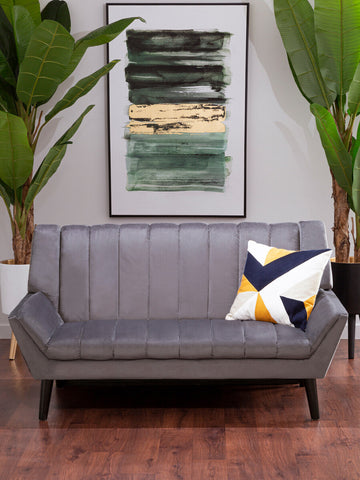 Two-seat grey sofa with a modern design, featuring vertical stitching on the backrest and tapered wooden legs. A decorative pillow with a geometric pattern in yellow, navy, and white is placed on one side. In the background, there is a large abstract painting and two tall green plants, set against a light-colored wall and wooden floor