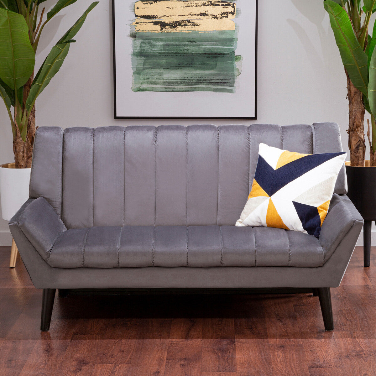 A stylish two-seat grey sofa featuring a modern design with vertical stitching on the backrest, complemented by a colorful geometric throw pillow. The sofa is positioned on a wooden floor, surrounded by indoor plants and a framed artwork in the background