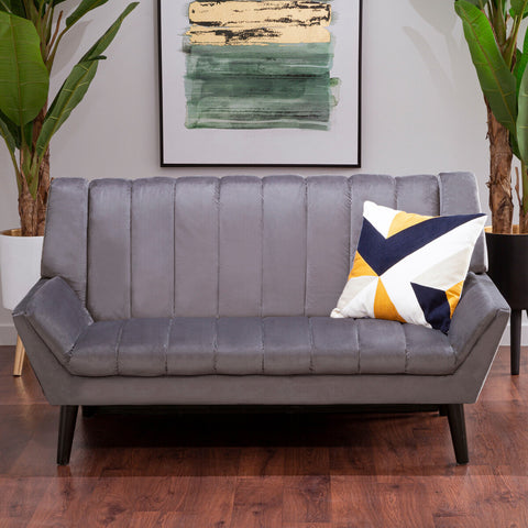 A stylish two-seat grey sofa featuring a modern design with vertical stitching on the backrest, complemented by a colorful geometric throw pillow. The sofa is positioned on a wooden floor, surrounded by indoor plants and a framed artwork in the background