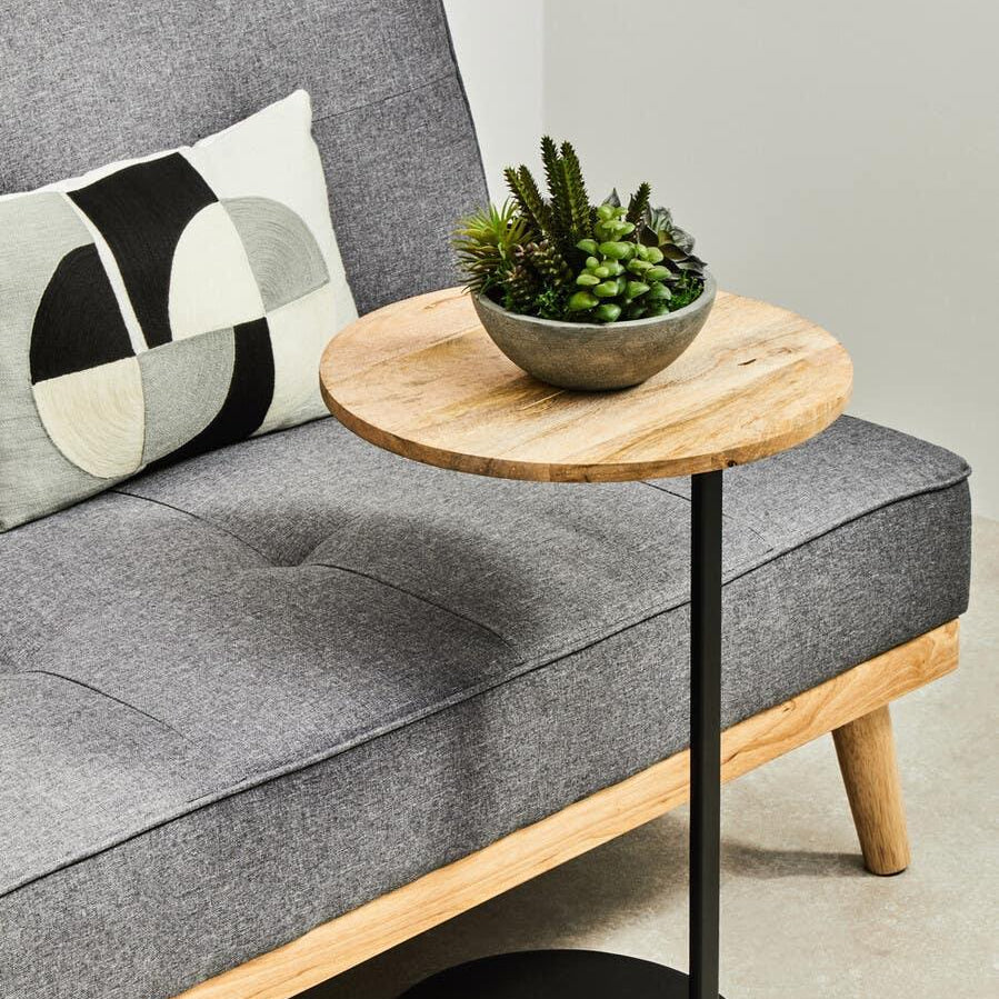 Round wooden side table with black metal base beside grey fabric sofa and cushion