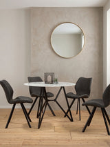 Round dining table with a marble effect top surrounded by six black chairs, set against a light-colored wall. A round mirror hangs above the table, and a small decorative centerpiece is placed on the table. The floor is made of light wood