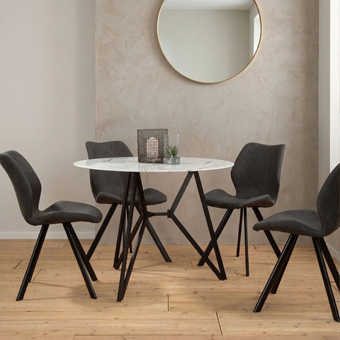 Round dining table with a marble effect top, surrounded by six modern black chairs with angled legs. The setting features a light-colored wall and a round mirror in the background, with a decorative centerpiece on the table. The floor is made of light wood