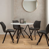 Round dining table with a marble effect top, surrounded by six modern black chairs with angled legs. The setting features a light-colored wall and a round mirror in the background, with a decorative centerpiece on the table. The floor is made of light wood