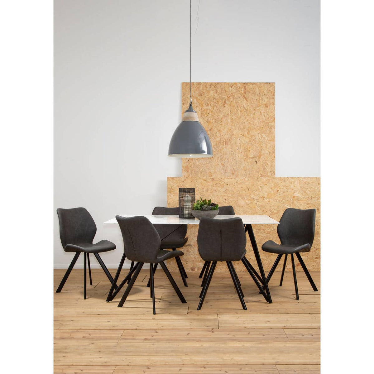 A modern dining set featuring a marble effect table surrounded by six stylish black chairs with wooden legs. The table is positioned on a light wooden floor, and a large pendant light hangs above, illuminating the space. A decorative plant and a few items are placed on the table, with a textured wall in the background