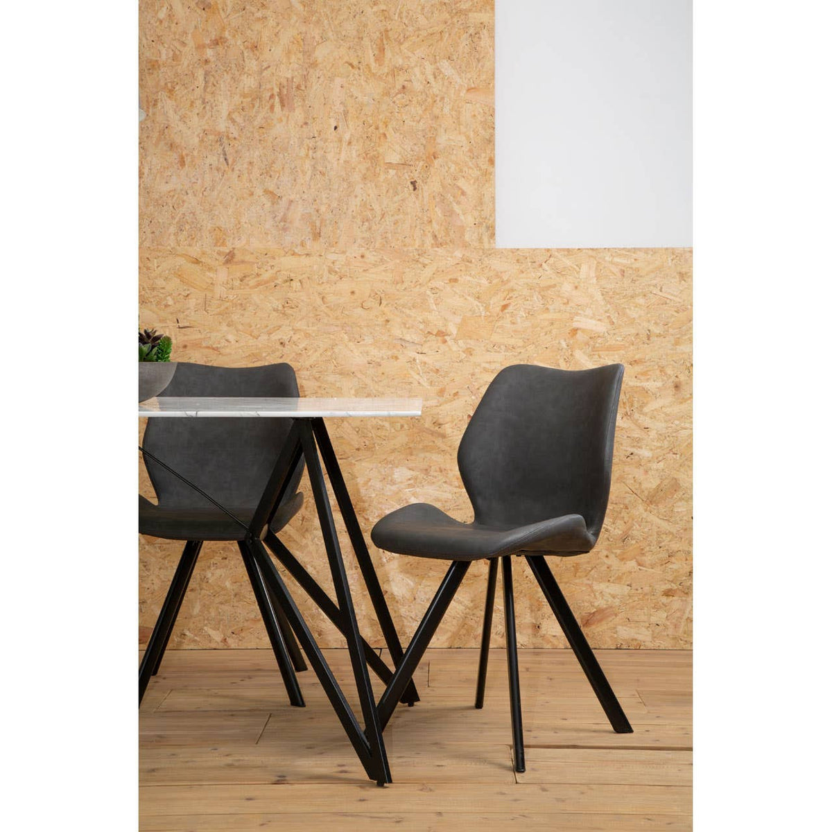 Weston Marble Effect Dining Table with six stylish gray chairs, featuring a sleek marble top and modern black metal legs, set against a textured wooden wall. The chairs have a contemporary design with curved backs and tapered legs, arranged around the table