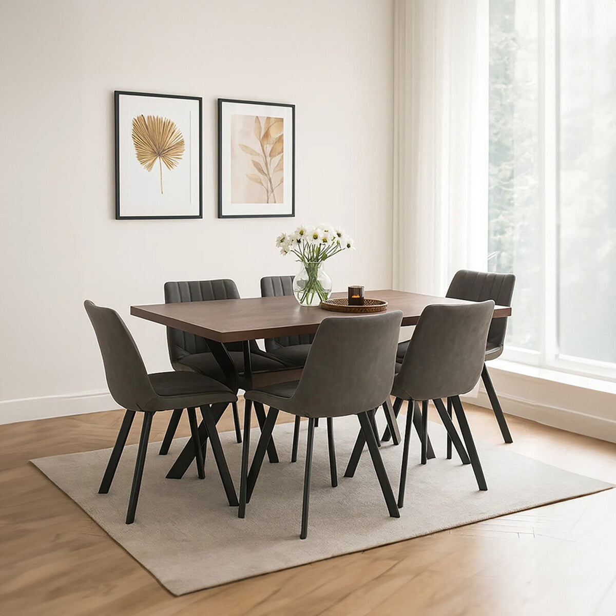 A rectangular wooden dining table surrounded by six upholstered chairs with dark legs, set in a bright room with large windows. A vase of flowers sits on the table, and two framed artworks are displayed on the wall. A light-colored area rug is positioned under the table and chairs, complementing the wooden floor