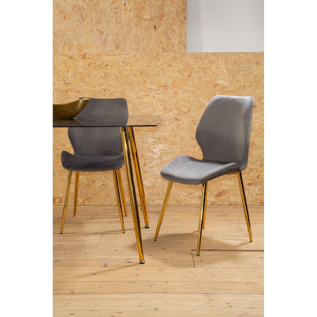 Smoked glass dining table with sleek, modern design featuring golden legs, accompanied by two upholstered chairs in a soft gray fabric with matching golden legs, set against a light wood backdrop