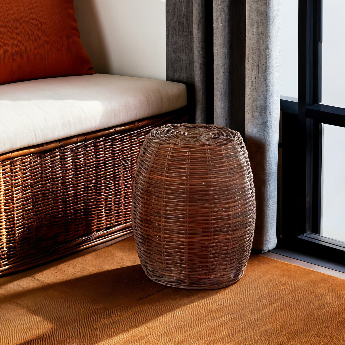 A natural croco rattan stool with a textured surface, positioned beside a woven bench in a well-lit room. The stool features a cylindrical shape and rich brown tones, complementing the wooden floor and the soft furnishings nearby
