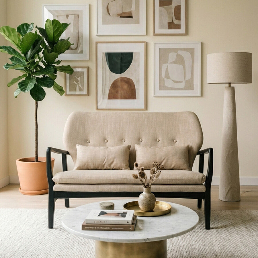 Beige two-seat sofa with a black wood frame, featuring two matching cushions, positioned in a cozy living room setting. A round marble coffee table sits in front of the sofa, surrounded by a light-colored rug. A potted plant and a floor lamp are visible in the background, along with framed artwork on the wall