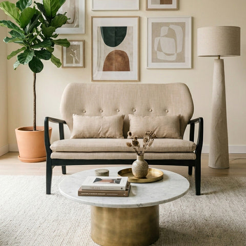 Beige two-seat sofa with a black wood frame, featuring tufted cushions and two decorative pillows, positioned in a cozy living room setting. A round coffee table with a marble top and a textured gold base sits in front of the sofa, surrounded by a light-colored rug. A tall floor lamp and a potted plant add to the decor, with framed artwork displayed on the wall behind the sofa