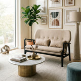 Beige two-seat sofa with a black wood frame, featuring tufted cushions and two decorative pillows, positioned in a bright living room with large windows. A round marble coffee table sits in front of the sofa, adorned with a decorative tray and a small plant. The background includes framed artwork on the walls and a tall floor lamp beside the sofa. A green potted plant adds a touch of nature to