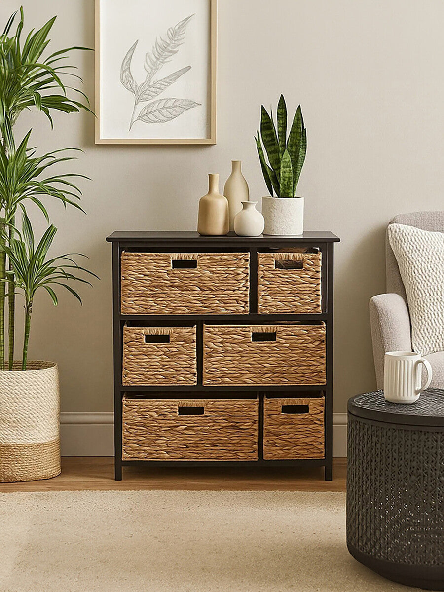 Black storage unit featuring six woven water hyacinth drawers with cut-out handles, positioned against a light-colored wall. A potted plant and decorative items are displayed on top of the unit, with a cozy armchair and a round side table visible in the foreground