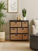 Black storage unit featuring six woven water hyacinth drawers with cut-out handles, positioned against a light-colored wall. A potted plant and decorative items are displayed on top of the unit, with a cozy armchair and a round side table visible in the foreground