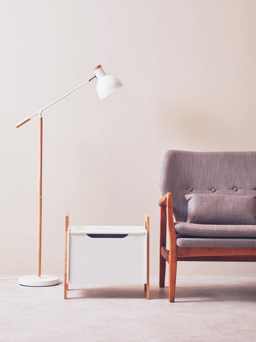 A white storage box with a black handle sits next to a modern gray sofa with a cushion, accompanied by a tall, minimalist floor lamp with a round shade, all set against a light-colored wall
