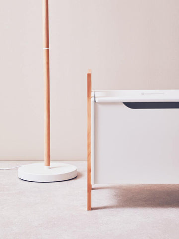 A white storage box with a sleek design featuring a single drawer, complemented by a minimalist floor lamp with a thin wooden pole and a round base. The background is a soft, neutral color, enhancing the modern aesthetic of the furniture