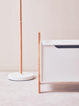 A white storage box with a sleek design featuring a single drawer, complemented by a minimalist floor lamp with a thin wooden pole and a round base. The background is a soft, neutral color, enhancing the modern aesthetic of the furniture