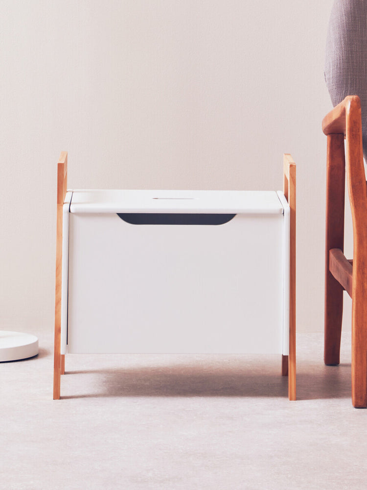 White storage box with a sleek design, featuring a dark handle and supported by light wooden legs. The box is positioned on a light-colored floor, with a soft, neutral background and a wooden chair partially visible beside it