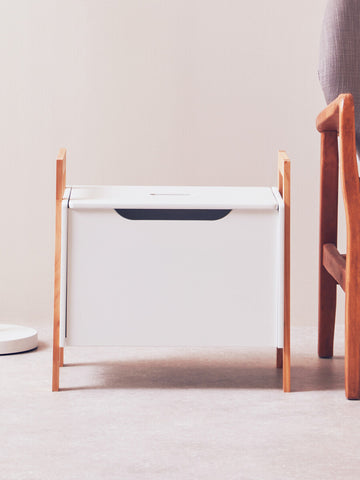 White storage box with a sleek design, featuring a dark handle and supported by light wooden legs. The box is positioned on a light-colored floor, with a soft, neutral background and a wooden chair partially visible beside it