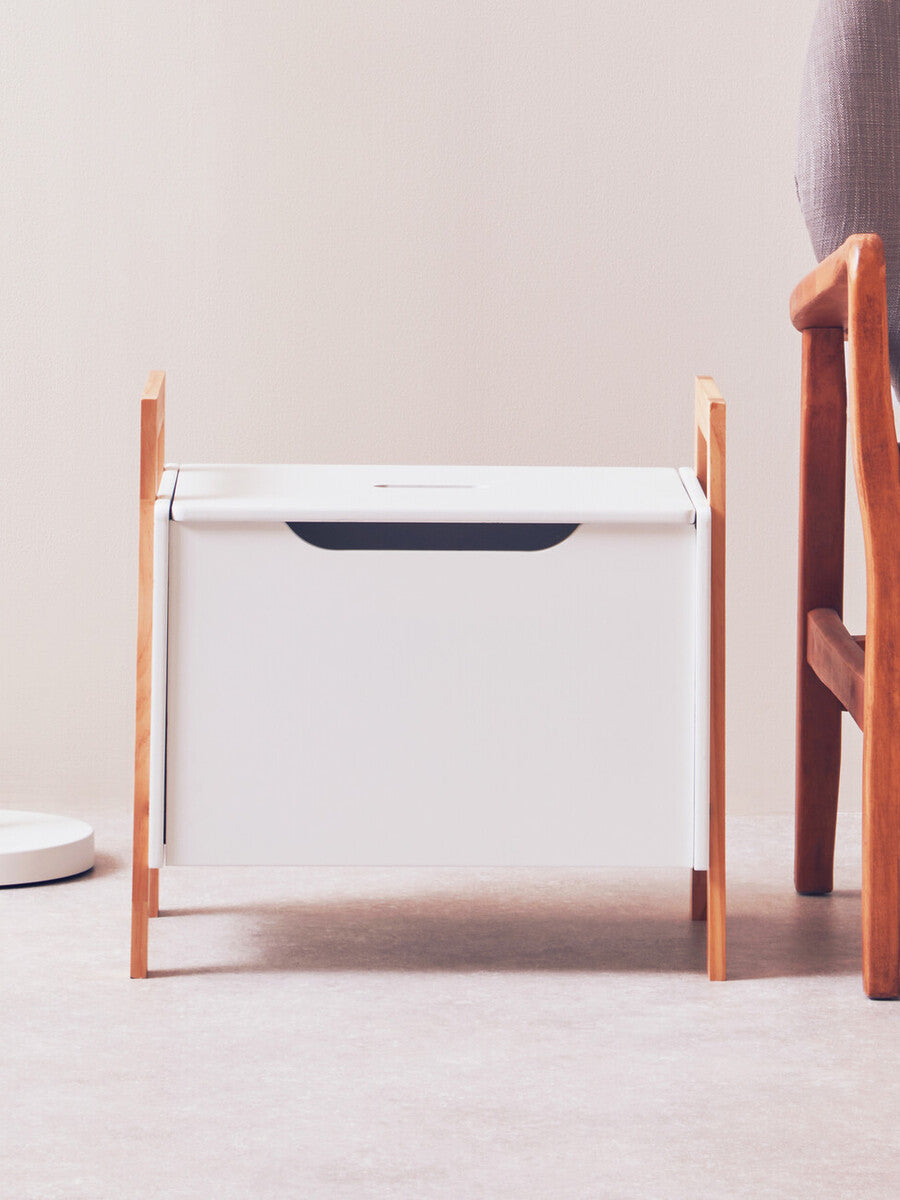 White storage box with a sleek design, featuring a dark handle and supported by light wooden legs. The box is positioned on a light-colored floor, with a soft, neutral background and a wooden chair partially visible beside it