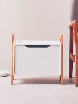 White storage box with a sleek design, featuring a dark handle and supported by light wooden legs. The box is positioned on a light-colored floor, with a soft, neutral background and a wooden chair partially visible beside it