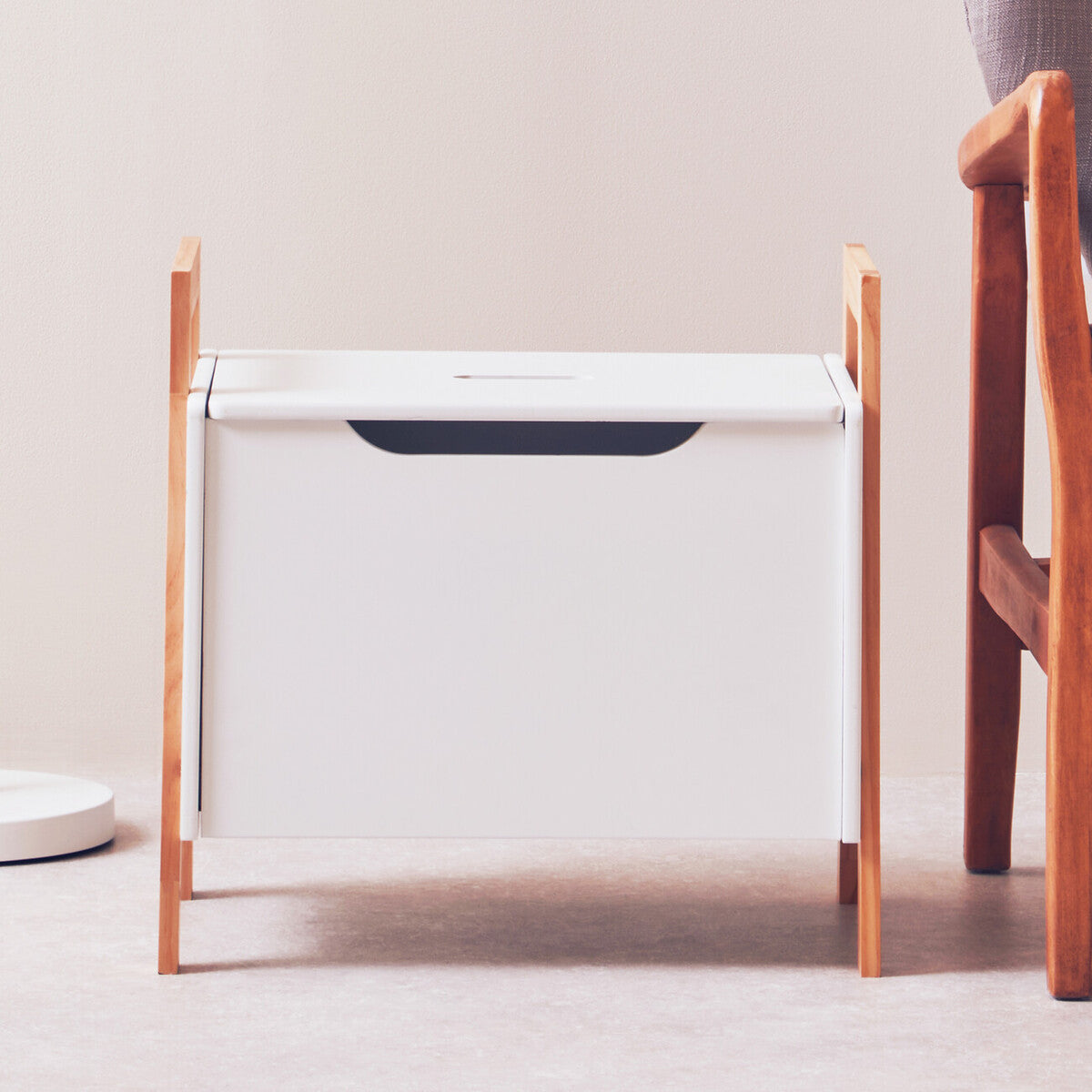 White storage box with a wooden frame, featuring a cut-out handle for easy access. The box is positioned on a light-colored floor, with a wooden chair partially visible in the background