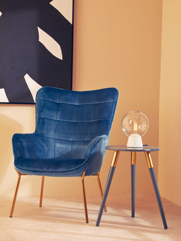 A dark grey side table with slender wooden legs stands next to a plush blue armchair. The table features a smooth, circular top and is adorned with a decorative glass lamp. The background showcases a warm, neutral wall with abstract art in black and white