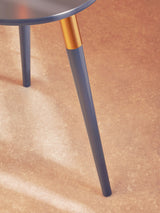 A dark grey side table with a round top and tapered legs, featuring a gold accent at the top of each leg, set against a light-colored floor