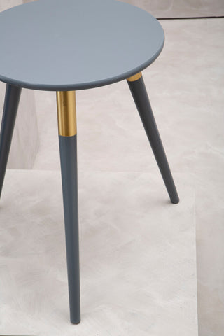 Round dark grey side table with three tapered legs, two of which have gold accents near the top. The table is placed on a light-colored floor, showcasing a minimalist design