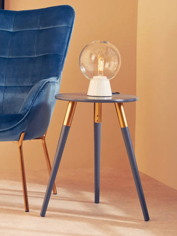 A dark grey side table with a smooth surface and tapered legs, featuring gold accents at the top of each leg. A stylish lamp with a round white base and a transparent globe sits on the table. The background includes a soft beige wall and a plush blue chair