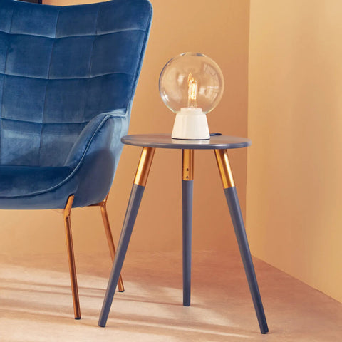 A dark grey side table with a smooth surface and three tapered legs, two of which are gold-colored. A stylish lamp with a round glass shade sits on top of the table, next to a blue upholstered chair against a warm-toned wall