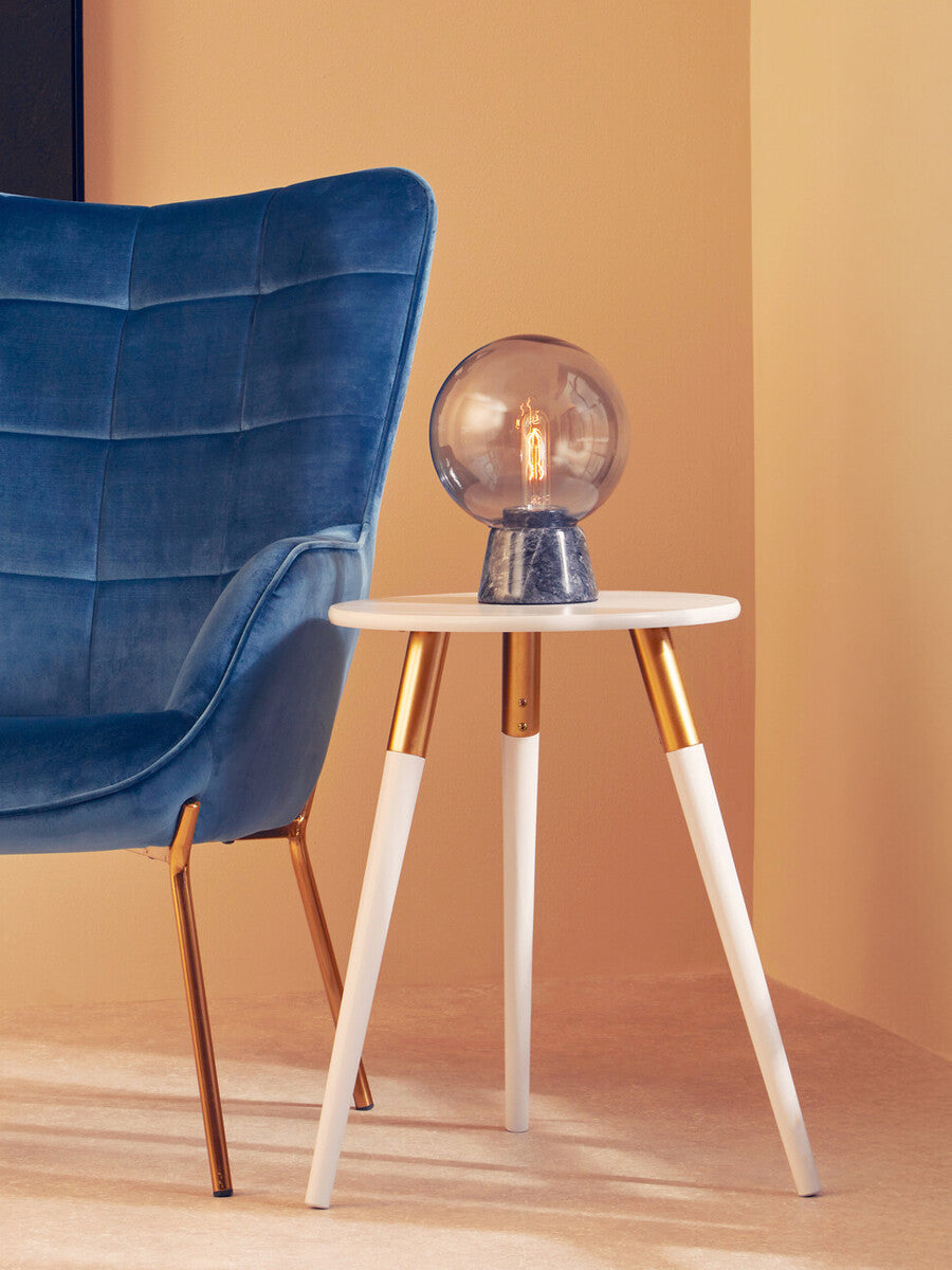 Light grey side table with a round top and four tapered wooden legs, positioned next to a blue upholstered chair. A decorative lamp with a transparent globe sits on the table, against a warm, neutral wall background