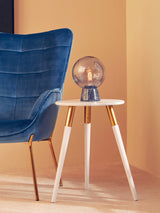 Light grey side table with a round top and four tapered wooden legs, positioned next to a blue upholstered chair. A decorative lamp with a transparent globe sits on the table, against a warm, neutral wall background