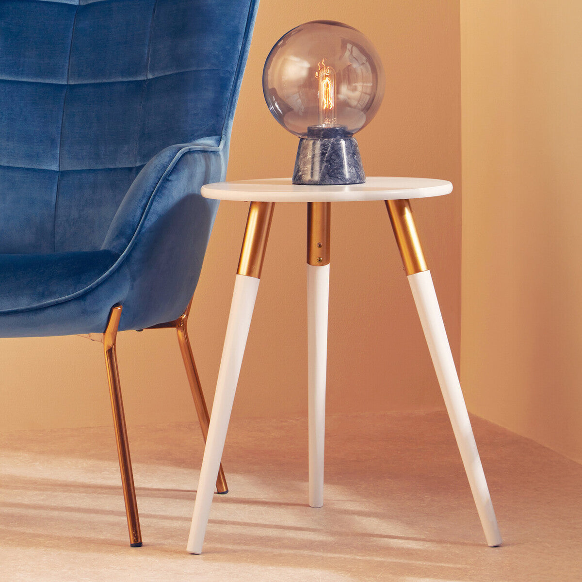 Light grey side table with a round top and three tapered legs, two of which are white and one is gold. A decorative globe lamp sits on the table, and a blue velvet chair is positioned nearby against a warm-toned wall