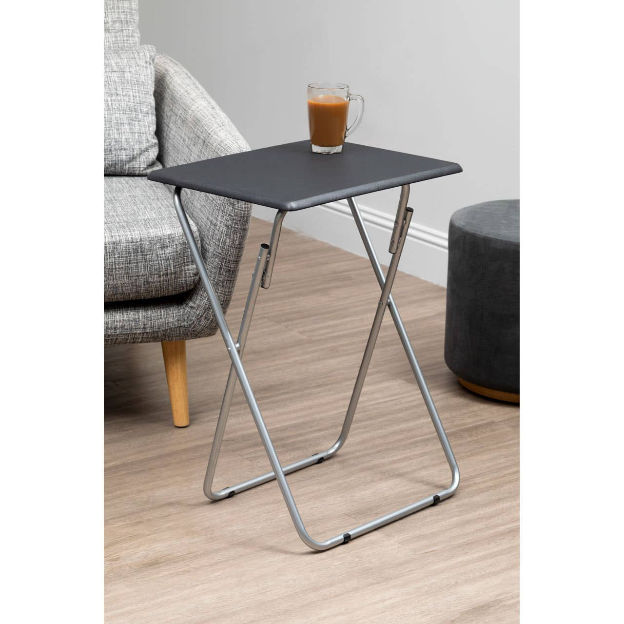 Folding table with a sand black top and a sleek metal frame, positioned next to a gray upholstered chair and a round gray ottoman, with a glass of beverage resting on the table surface. The floor features light wooden planks