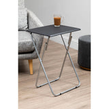Folding table with a sand black top and a sleek metal frame, positioned next to a gray upholstered chair and a round gray ottoman, with a glass of beverage resting on the table surface. The floor features light wooden planks