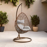 A brown rattan hanging chair with a curved frame and a circular base, featuring plush white cushions inside. The chair is suspended from a tall, arched support, set against a neutral-colored wall with potted greenery in the background