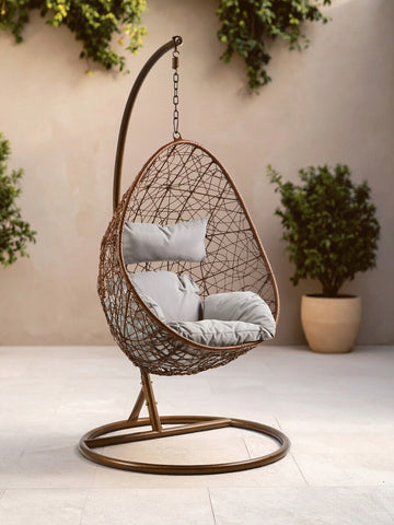 A brown rattan hanging chair with a curved design, featuring a plush white cushion, suspended from a sleek metal stand. The chair is positioned on a light-colored floor, surrounded by potted greenery, creating a cozy and inviting atmosphere