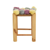 A small wooden stool with a natural finish, featuring a top adorned with colorful pom poms in shades of grey, beige, pink, and yellow. The stool has a simple, minimalist design with two horizontal support bars