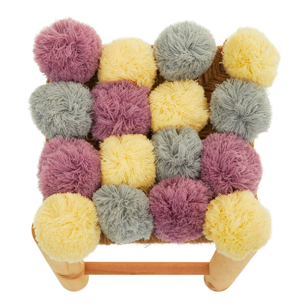 A square stool featuring a colorful arrangement of pom poms in shades of grey, beige, pink, and yellow, with a light wooden frame. The pom poms are plush and textured, creating a playful and vibrant design