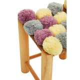 A stool featuring a wooden frame with a seat adorned with colorful pom poms in shades of grey, beige, pink, and yellow, creating a playful and textured design