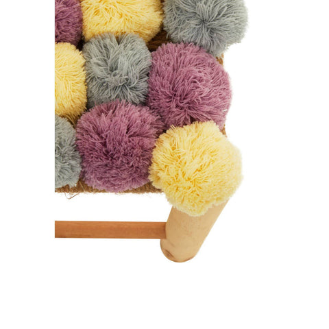 A close-up view of a stool featuring a colorful arrangement of pom poms in shades of grey, beige, pink, and yellow, with a wooden base and legs. The pom poms are fluffy and textured, creating a playful and vibrant look