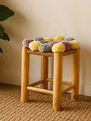 A wooden stool with a light natural finish, featuring a padded top adorned with grey, beige, pink, and yellow pom poms. The stool has four sturdy legs and is placed on a textured woven rug, with a plant visible in the background