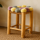 A wooden stool with a natural finish featuring a top adorned with colorful pom poms in shades of grey, beige, pink, and yellow. The stool has four sturdy legs and is placed on a textured woven rug