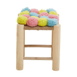 A stool with a wooden frame featuring a colorful top adorned with pink, blue, and yellow pom poms. The stool has a simple design with three horizontal support bars on the sides, showcasing a playful and vibrant aesthetic suitable for home decor