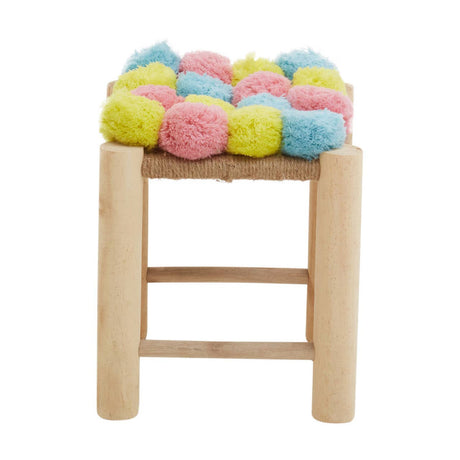 A stool with a wooden frame featuring a colorful top adorned with pink, blue, and yellow pom poms. The stool has a simple design with three horizontal support bars on the sides, showcasing a playful and vibrant aesthetic suitable for home decor