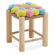 A wooden stool with four light-colored legs and a square top adorned with colorful pom poms in pink, blue, and yellow. The pom poms are arranged in a playful pattern, adding a vibrant touch to the design