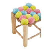 A small stool featuring a colorful pom pom seat with pink, blue, and yellow pom poms arranged in a playful pattern, supported by light wooden legs